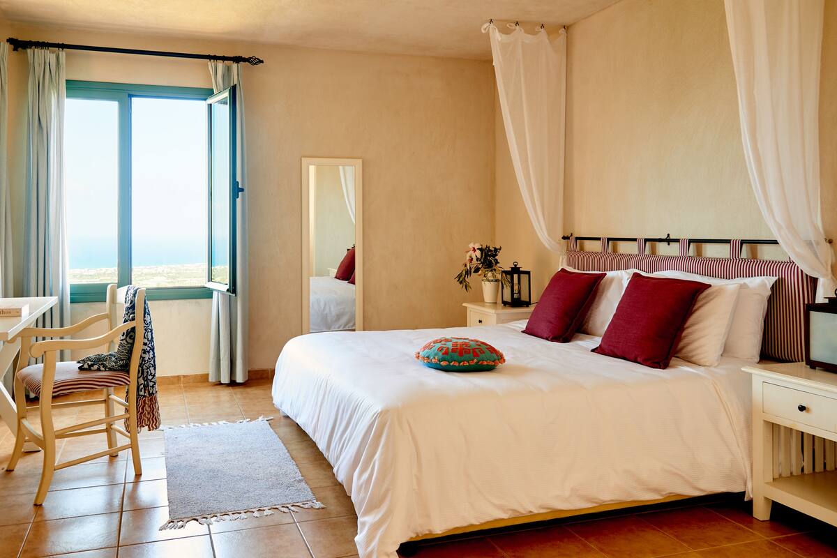 A bedroom with a bed, a bedside table, a chair, a window with curtains, and a view of the sea.
