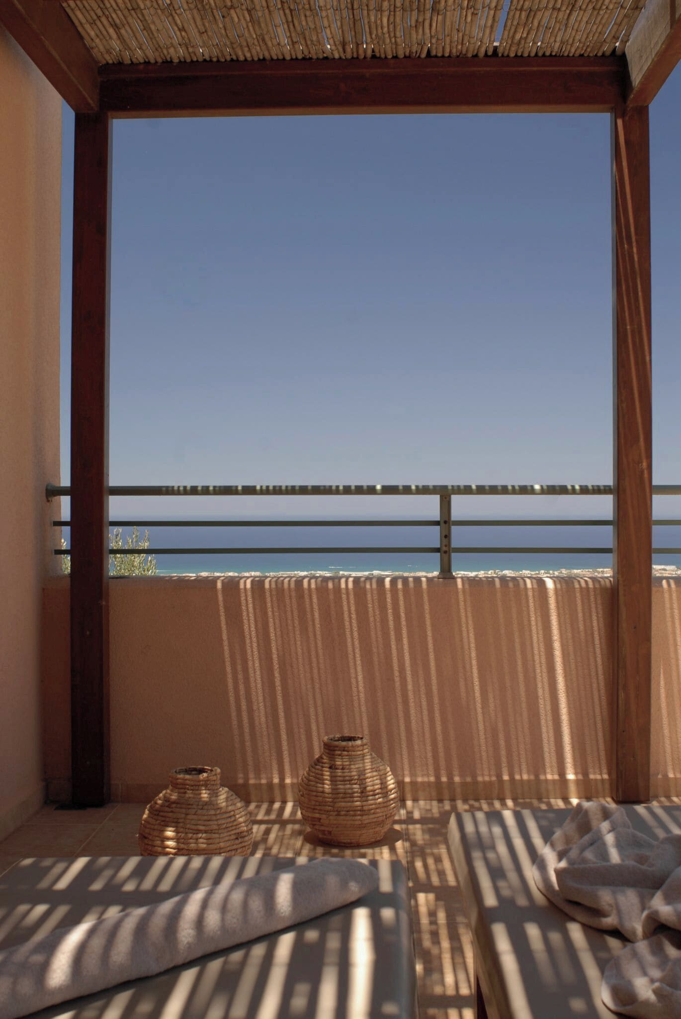 A balcony with a view of the sea, featuring a wooden pergola, a metal railing, and two woven baskets.