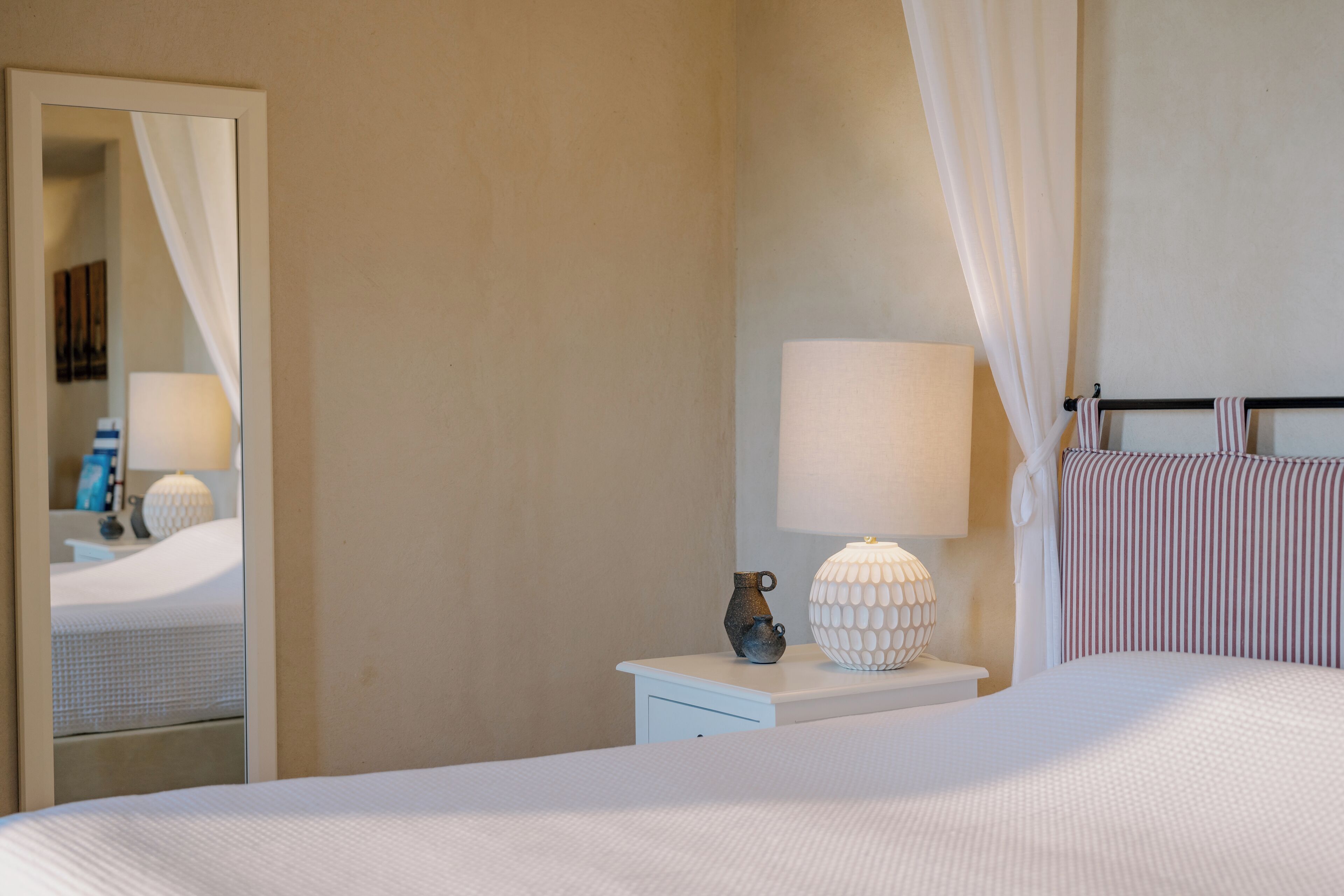 A neatly made bed with a white comforter, a bedside table with a lamp and a vase, and a mirror on the wall.