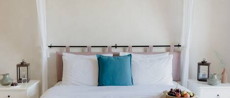 Una cama bien hecha con ropa de cama blanca y una sola almohada azul, flanqueada por almohadas a rayas. Un cuenco de madera con frutas está sobre la cama.