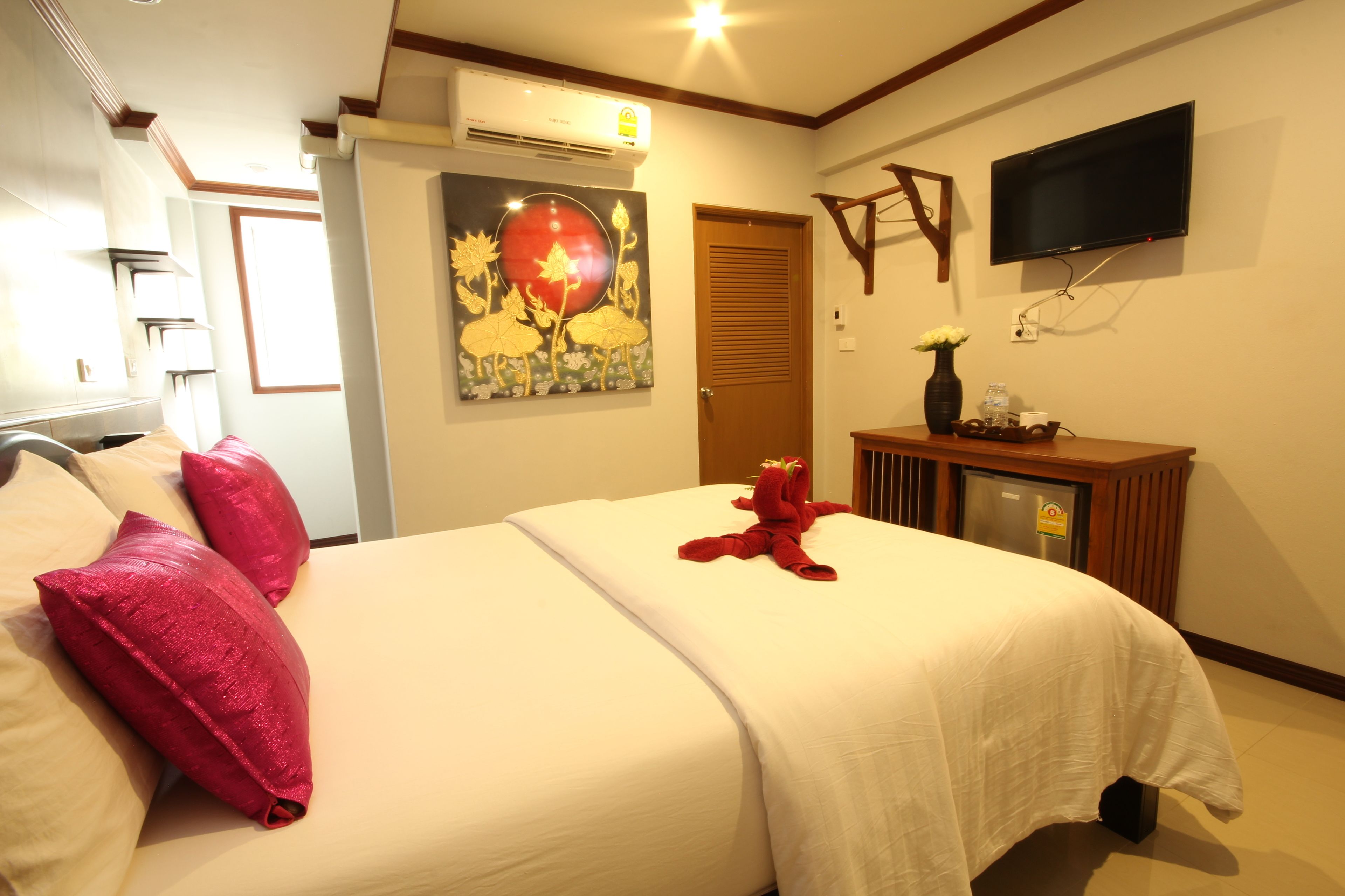 A hotel room with a bed, pillows, a painting, a TV, and a desk with a vase and a cup.