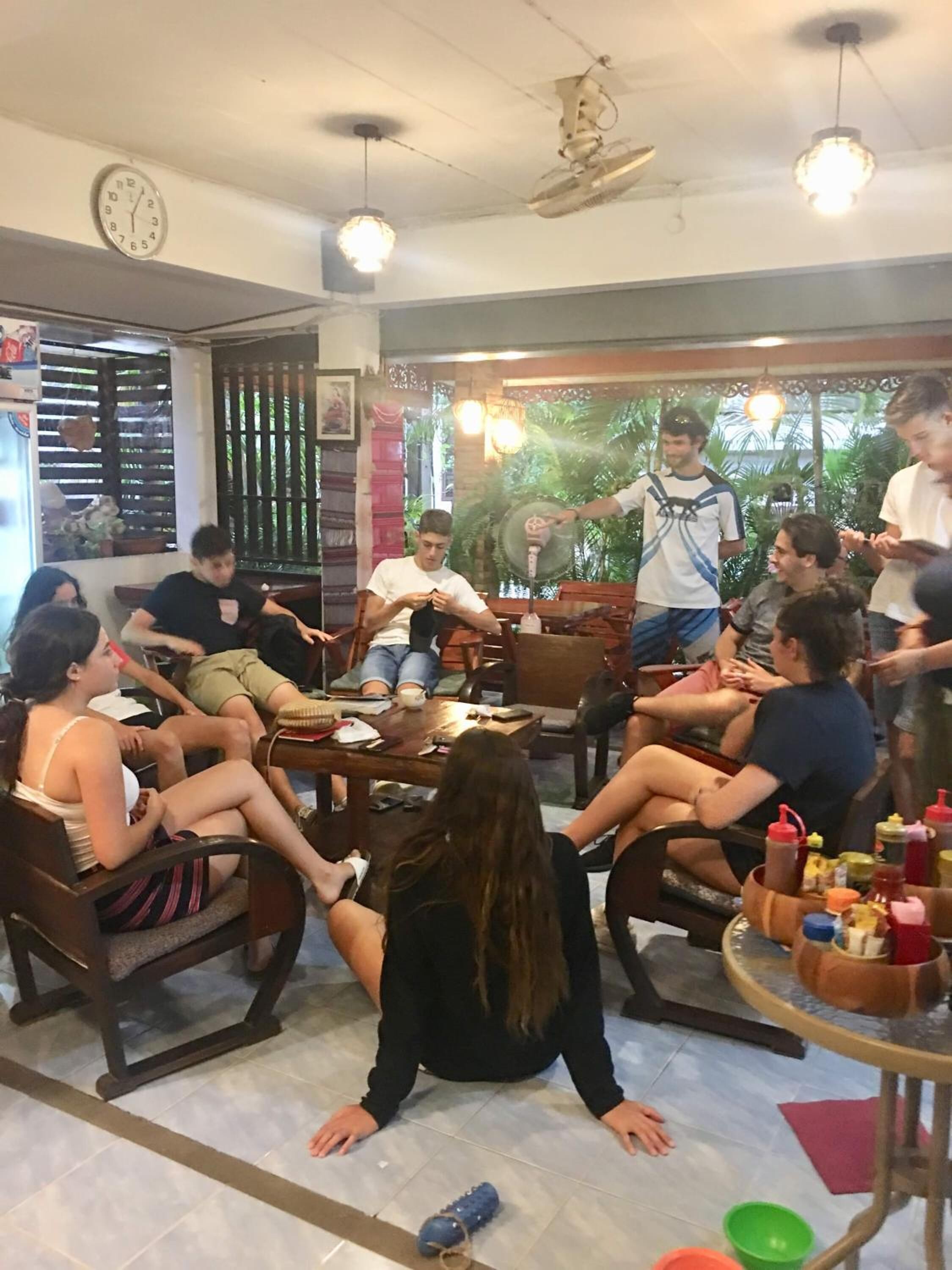 Un groupe de personnes est assis autour d’une table dans une pièce où l’on peut voir un ventilateur de plafond, une horloge et une fenêtre avec des stores.