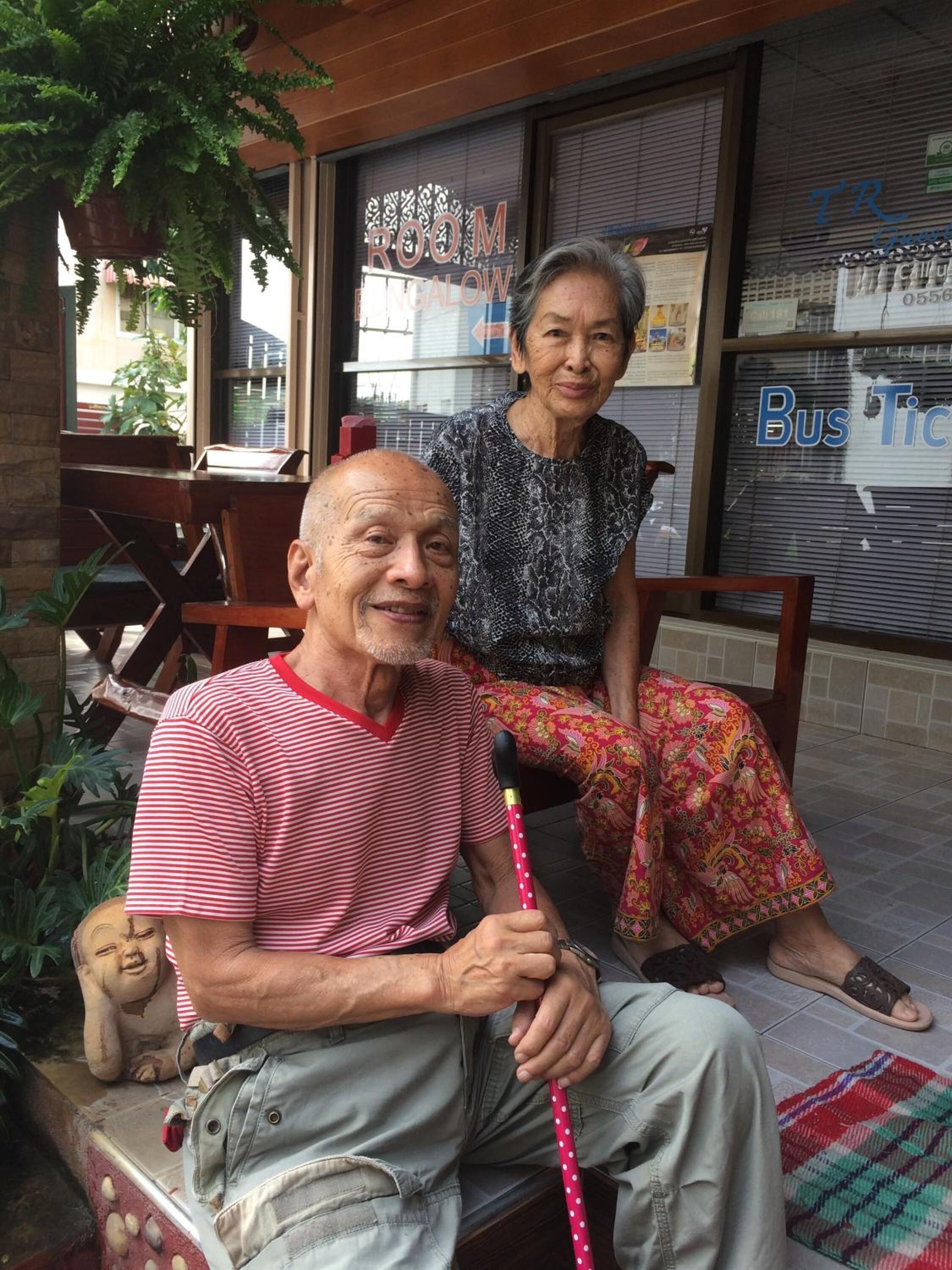 Una pareja de edad avanzada sentada en un banco frente a un edificio con un cartel que dice 'ROOM BENGALOW'.
