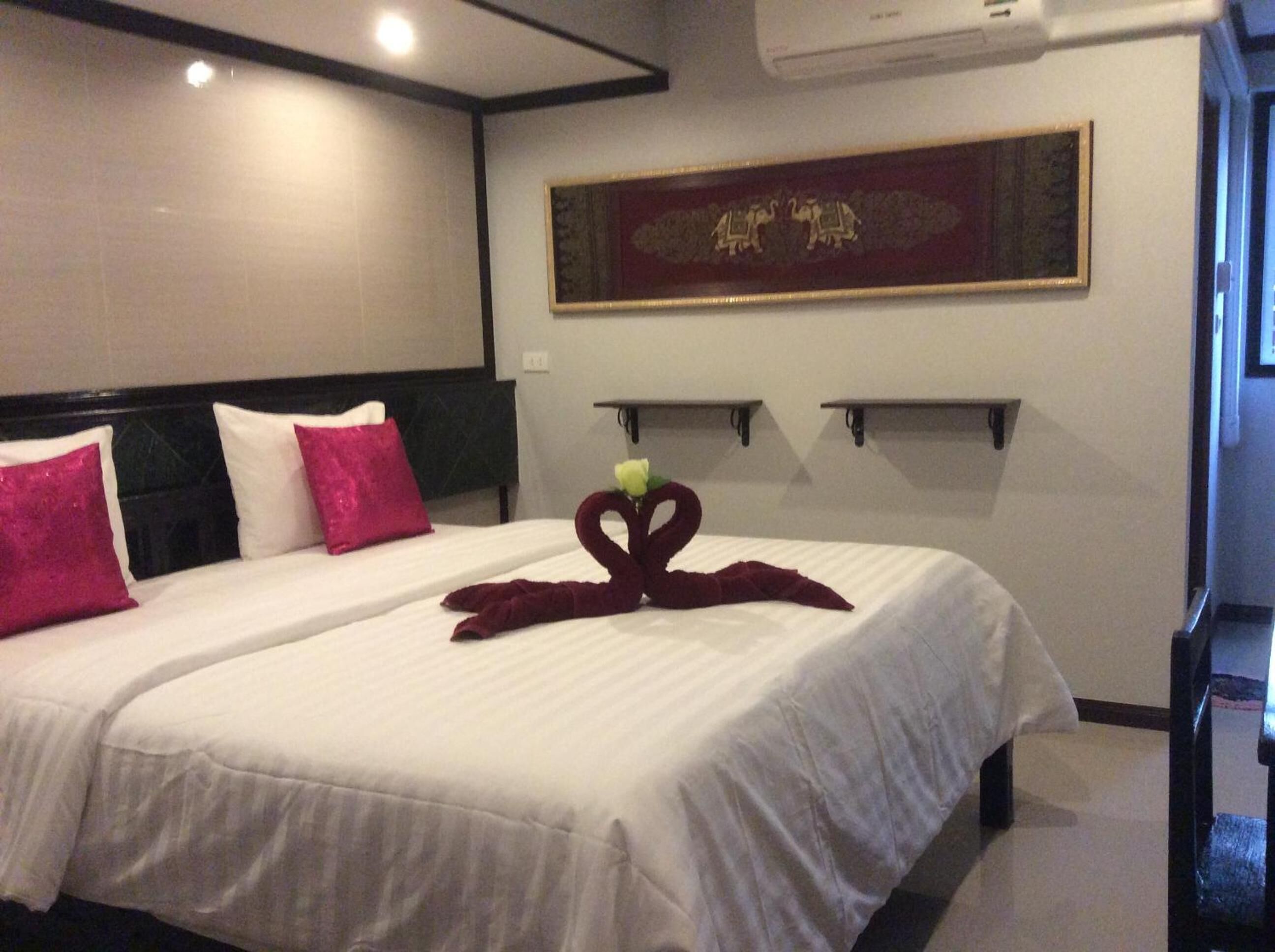 A hotel room with two beds, each with white bedding and pink pillows. There is a decorative swan towel on the bed in the foreground. The room has a modern design with a dark headboard and a wall-mounted air conditioner.