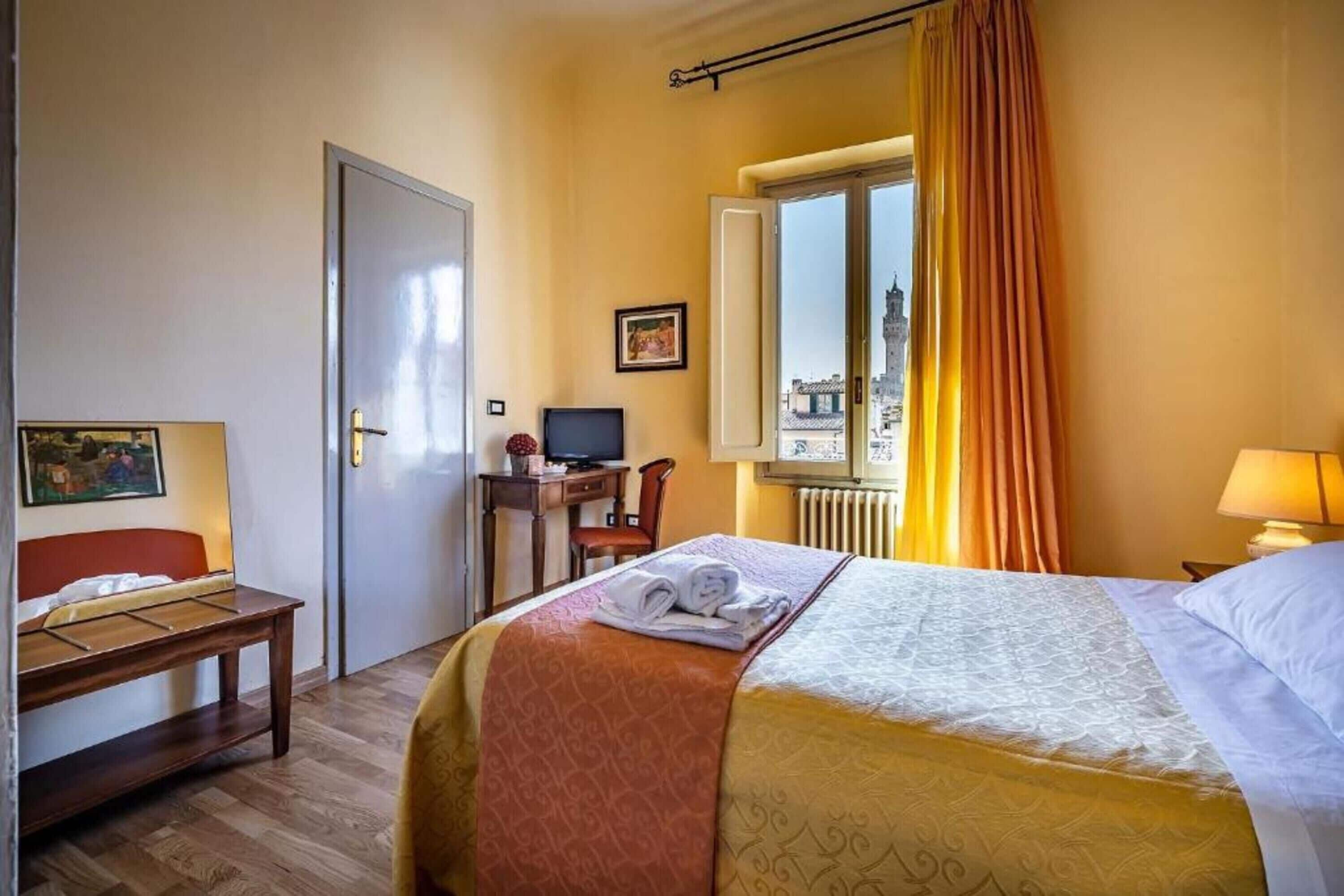 A hotel room with a bed, a desk with a computer, a chair, and a window with curtains.