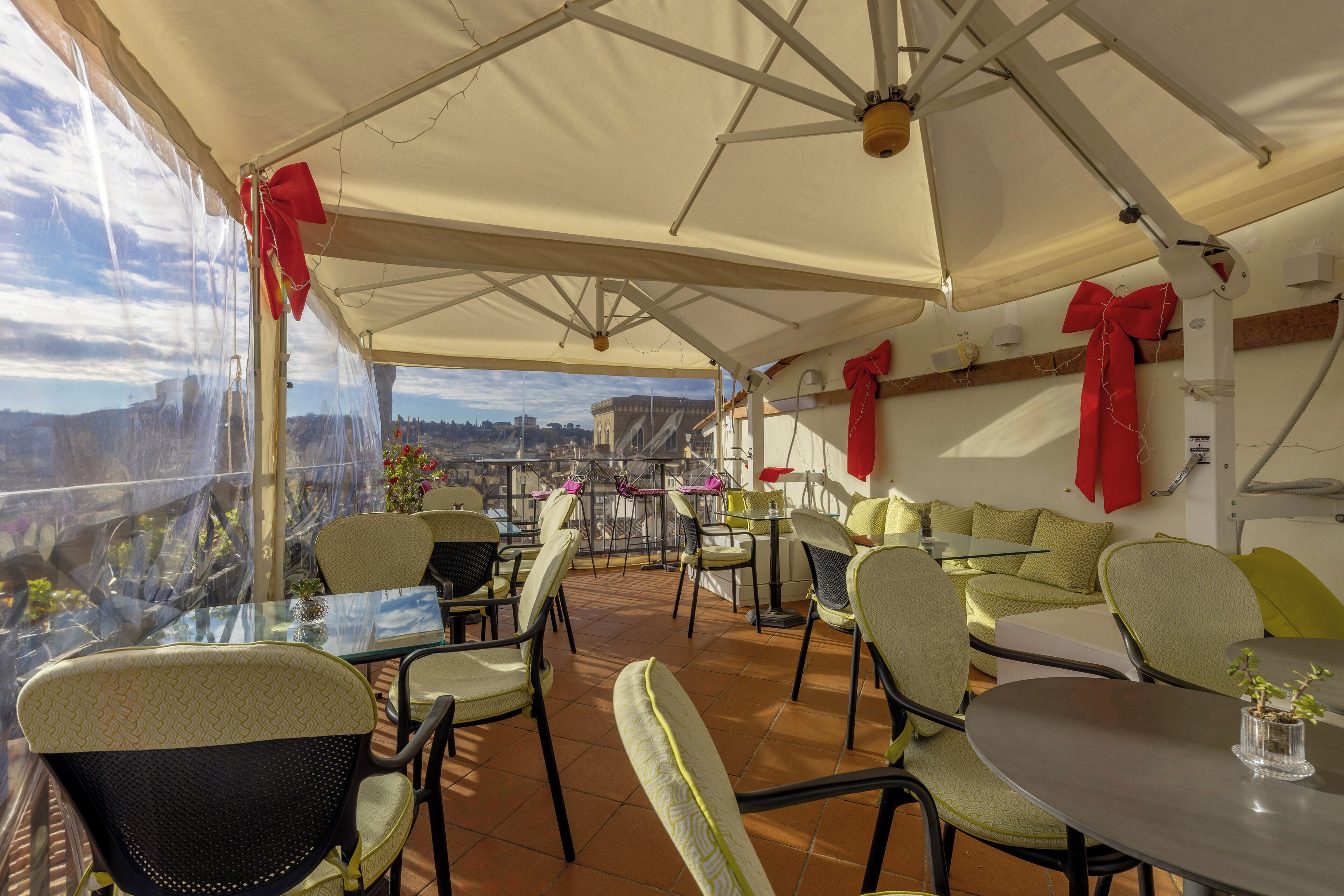 An outdoor terrace with tables and chairs, red bows on the ceiling, and a view of the city.