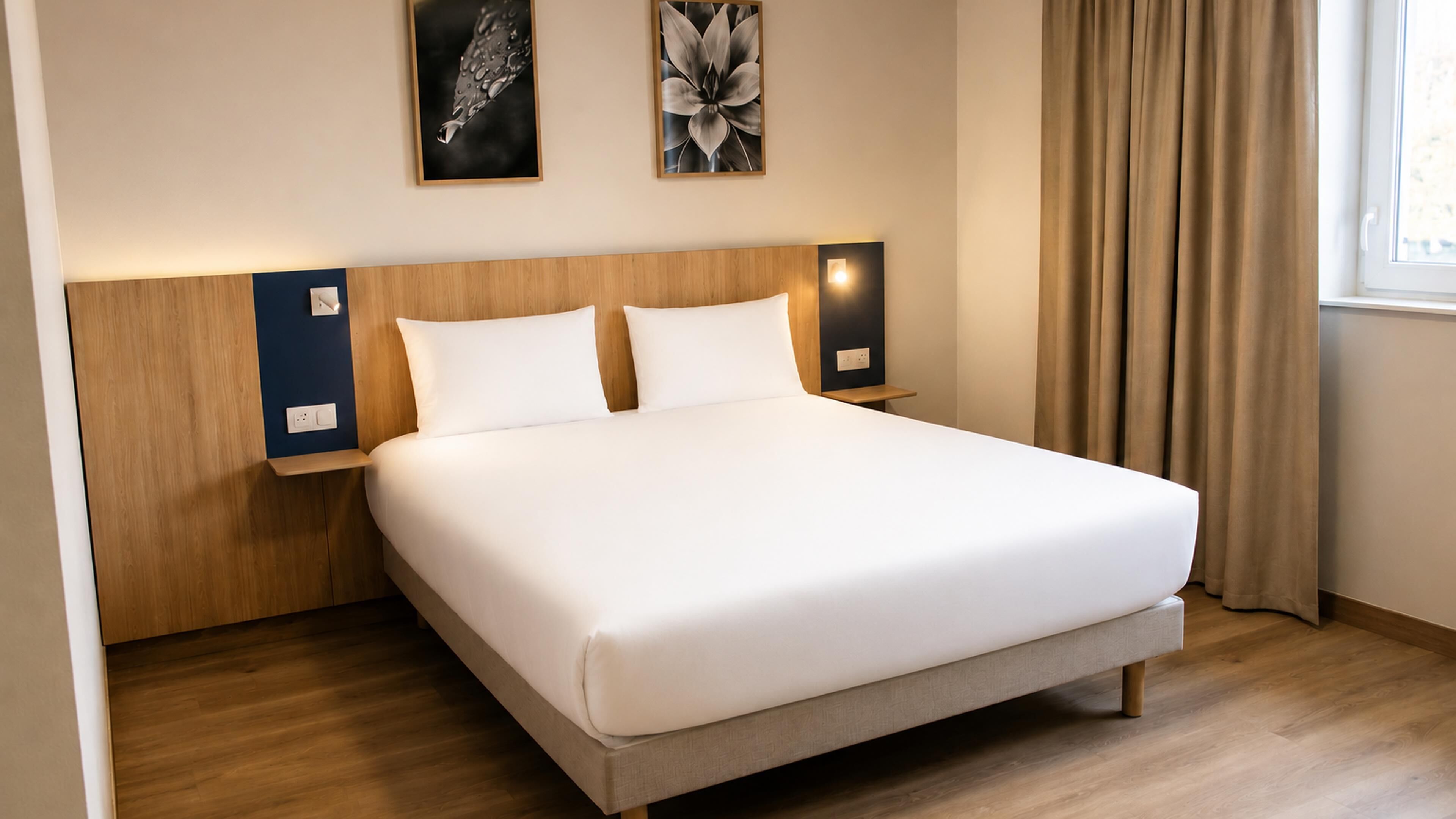 Une chambre d’hôtel avec un grand lit, une tête de lit en bois, deux tables de chevet fixées au mur et une fenêtre avec des rideaux.