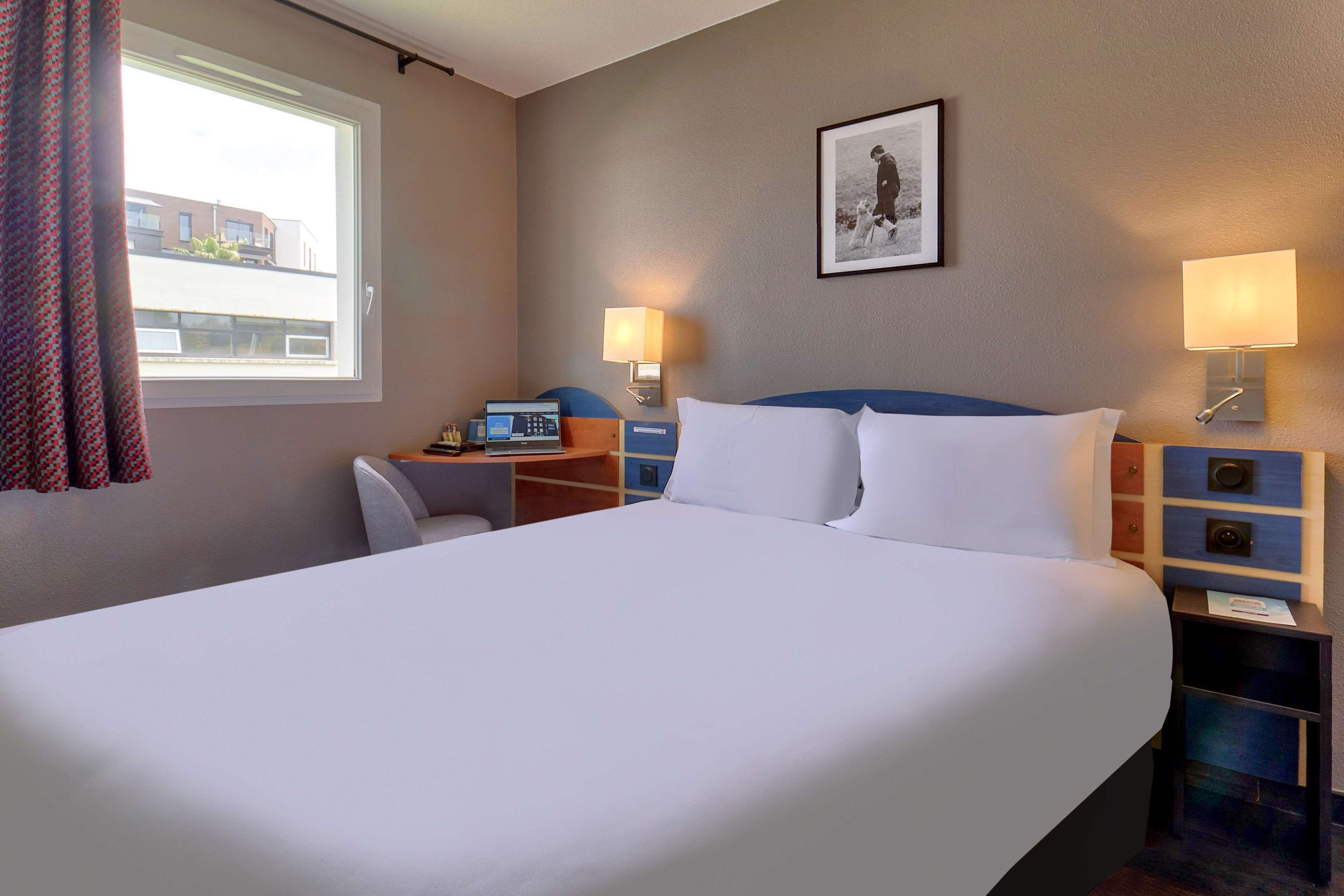 Quarto de hotel com uma cama grande, uma escrivaninha com um computador, duas luminárias de cabeceira e um quadro na parede.