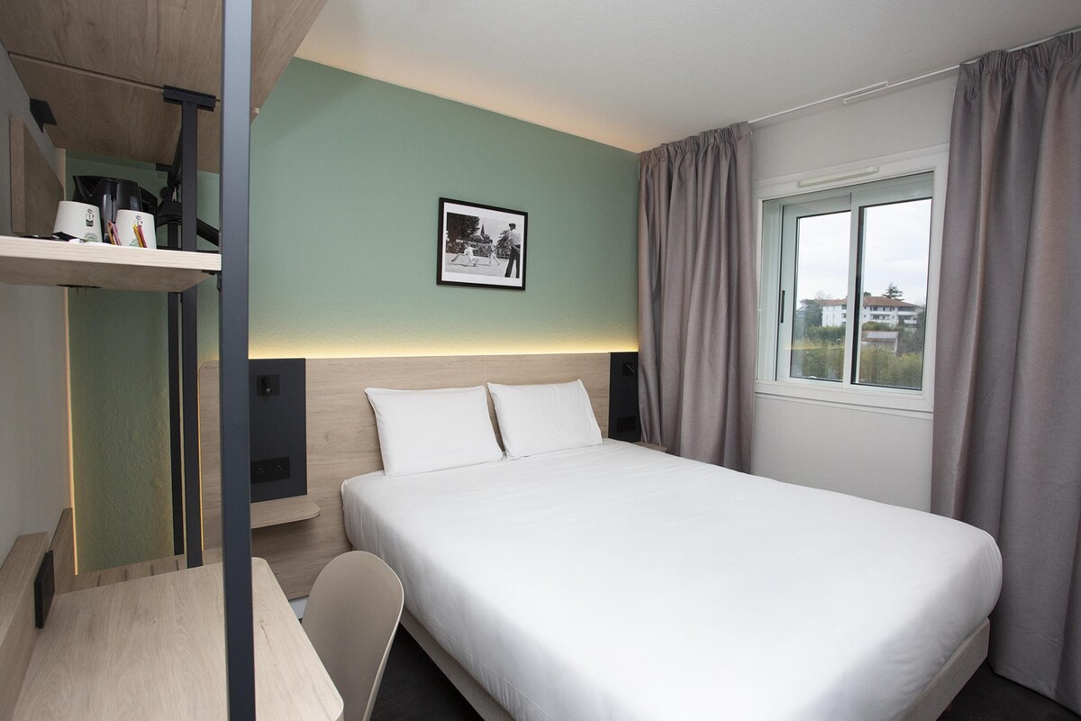 Habitación de hotel con cama, escritorio, silla, un cuadro en la pared y ventana con cortinas.