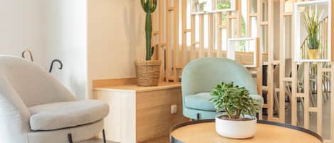 A modern living room with a grey armchair, a teal armchair, a round wooden coffee table with a potted plant, and a wooden partition with shelves.
