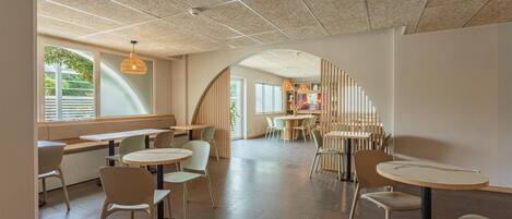 Una cafetería moderna con mesas y sillas redondas, techo inclinado y grandes ventanales.