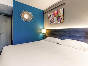 A hotel room with a blue accent wall, a sunburst mirror, a colorful artwork, a bed with white linens, and a nightstand with a lamp. - Greet Hotel Bordeaux Floirac Arena (Floirac)