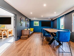 Een moderne kamer met houten vloer, een grote tafel met stoelen, een bank met kussens, en een televisie aan de muur.