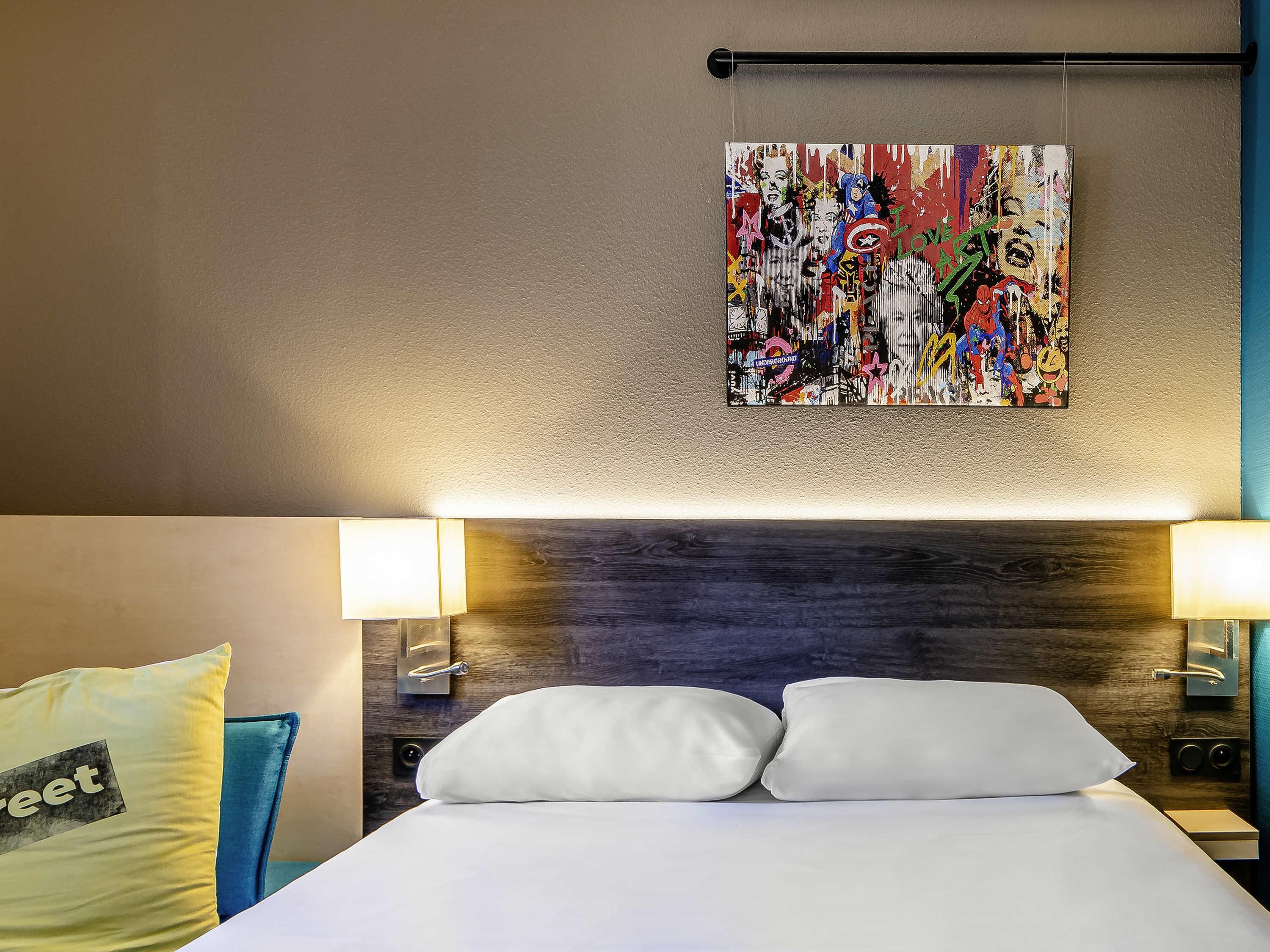A hotel room with a bed, white pillows, a colorful abstract painting on the wall, and two wall-mounted lamps.