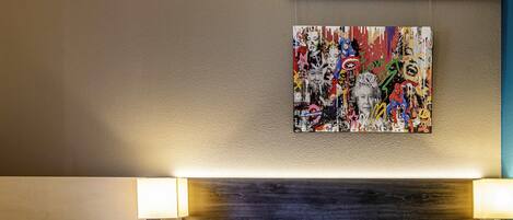 A hotel room with a bed, white pillows, a colorful abstract painting on the wall, and two wall-mounted lamps.