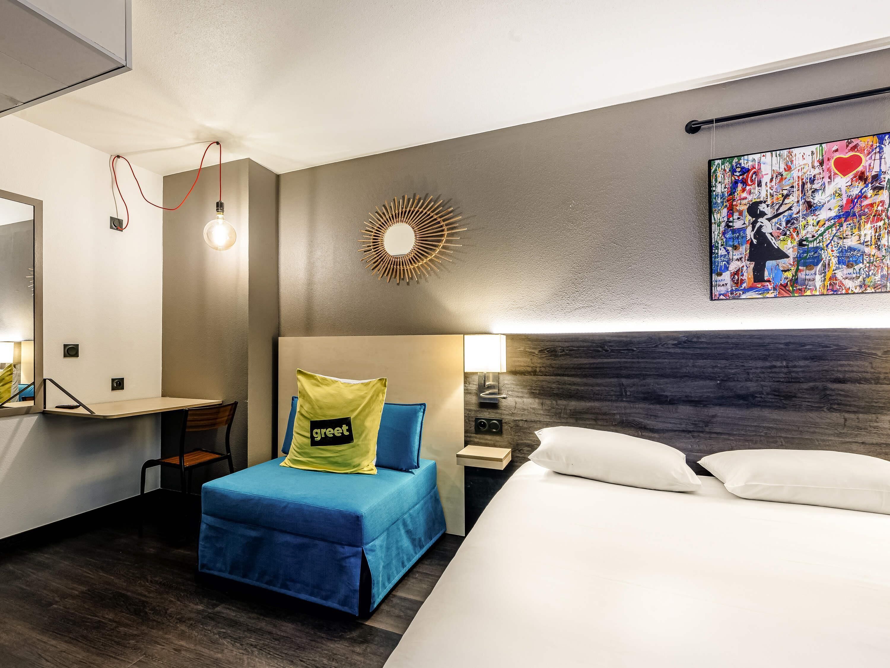 Habitación de hotel con cama, escritorio, silla, espejo, reloj de pared, mesita de noche con lámpara y un cuadro abstracto de colores.
