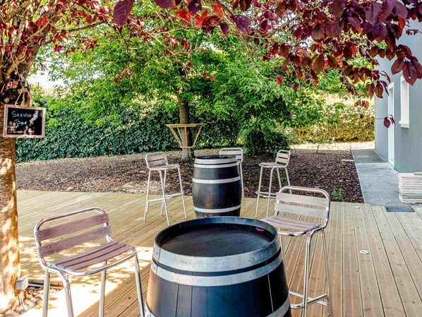 Un espace extérieur aménagé avec des chaises en métal et un grand tonneau, entouré de verdure et doté d’une terrasse en bois.