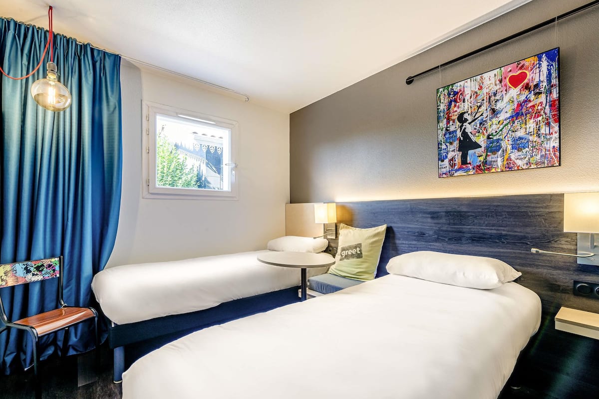 Ein Hotelzimmer mit zwei Betten, einem Fenster, einem Gemälde und einem Sessel.