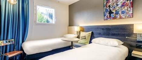 Habitación de hotel con dos camas, una mesita, una silla, un ventanal con cortinas, un cuadro en la pared y una lámpara.