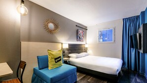 Une chambre d’hôtel avec un lit, un bureau, une chaise, un canapé bleu, un téléviseur fixé au mur et une fenêtre avec des rideaux.