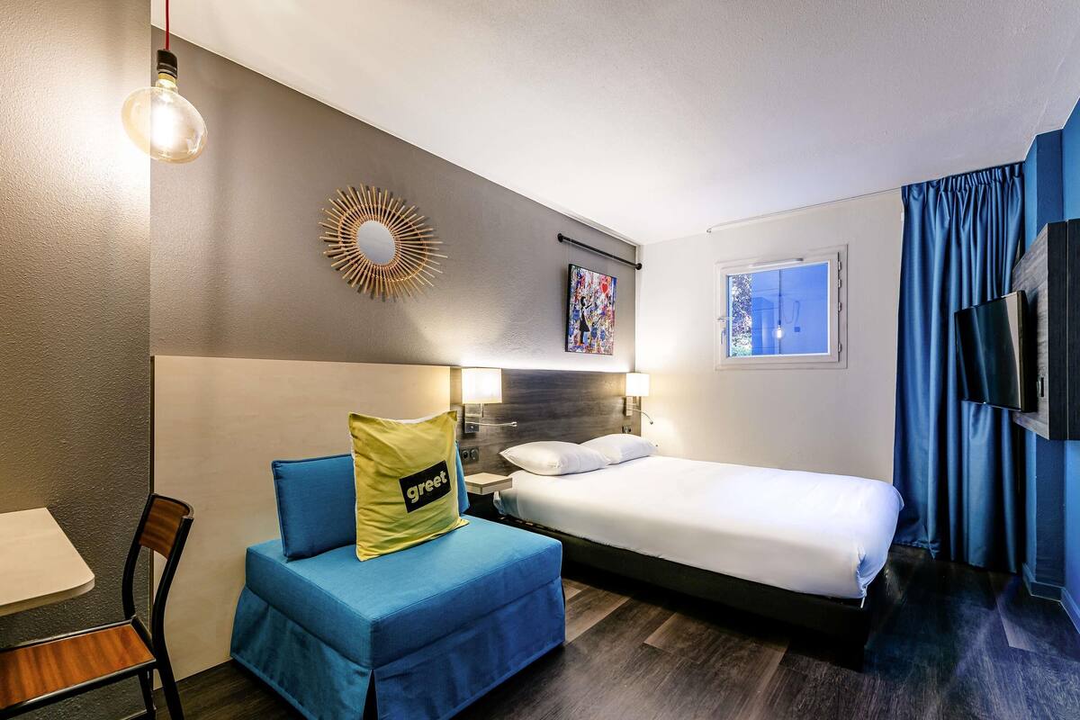 Ein Hotelzimmer mit Bett, Schreibtisch, Stuhl, blauem Sofa, Wandfernseher und einem Fenster mit Vorhängen.