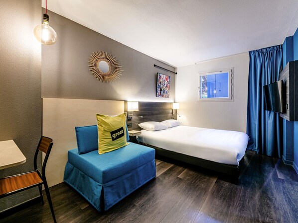 Ein Hotelzimmer mit Bett, Schreibtisch, Stuhl, blauem Sofa, Wandfernseher und einem Fenster mit Vorhängen.