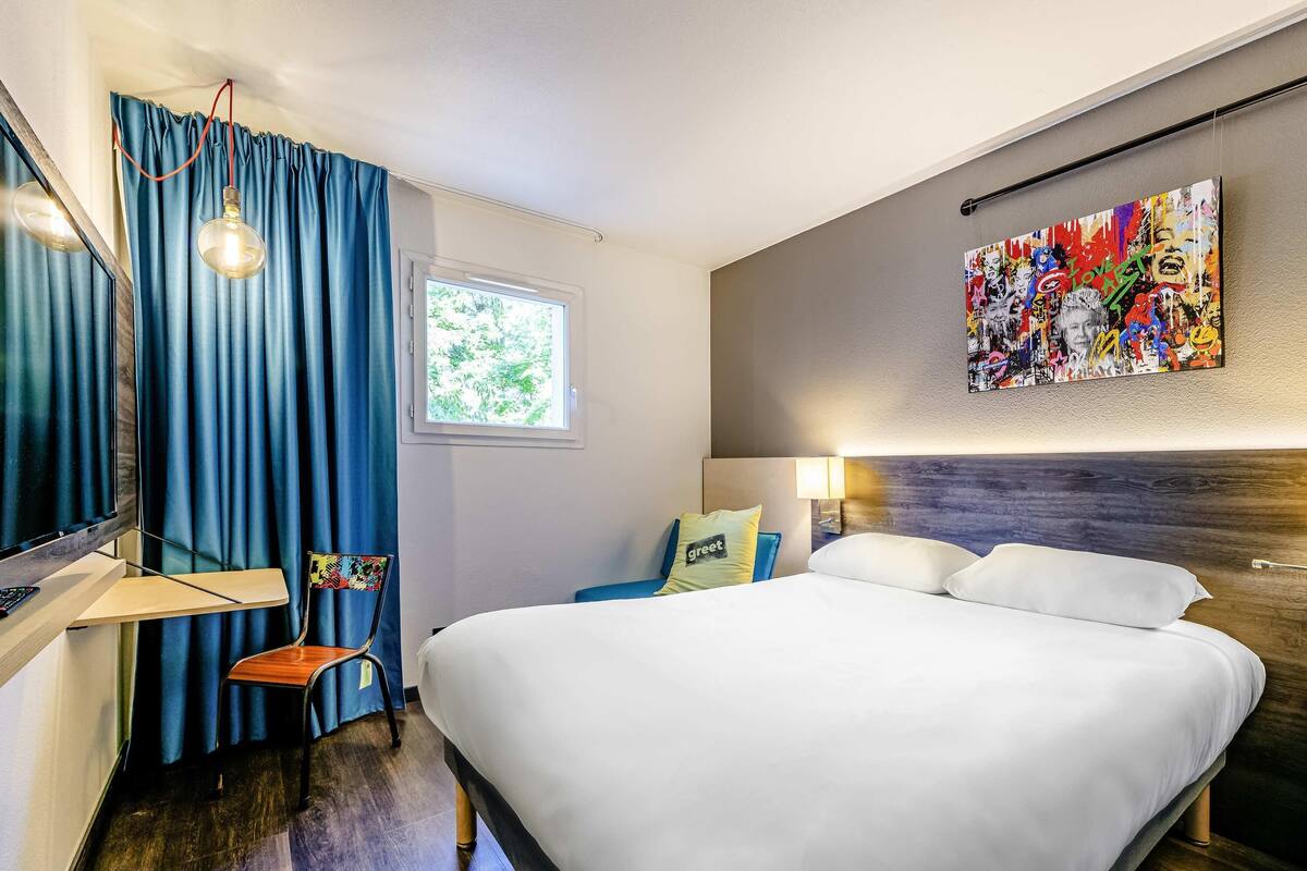 Ein Hotelzimmer mit Bett, Schreibtisch, Stuhl, Fernseher, Fenster und einem Wandbild.