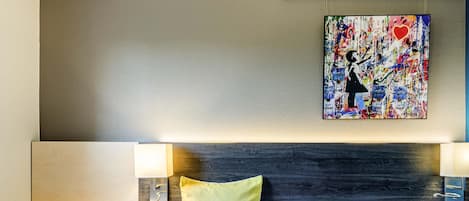 A hotel room with two beds, a round table, a headboard with a pillow, and a colorful abstract painting on the wall.
