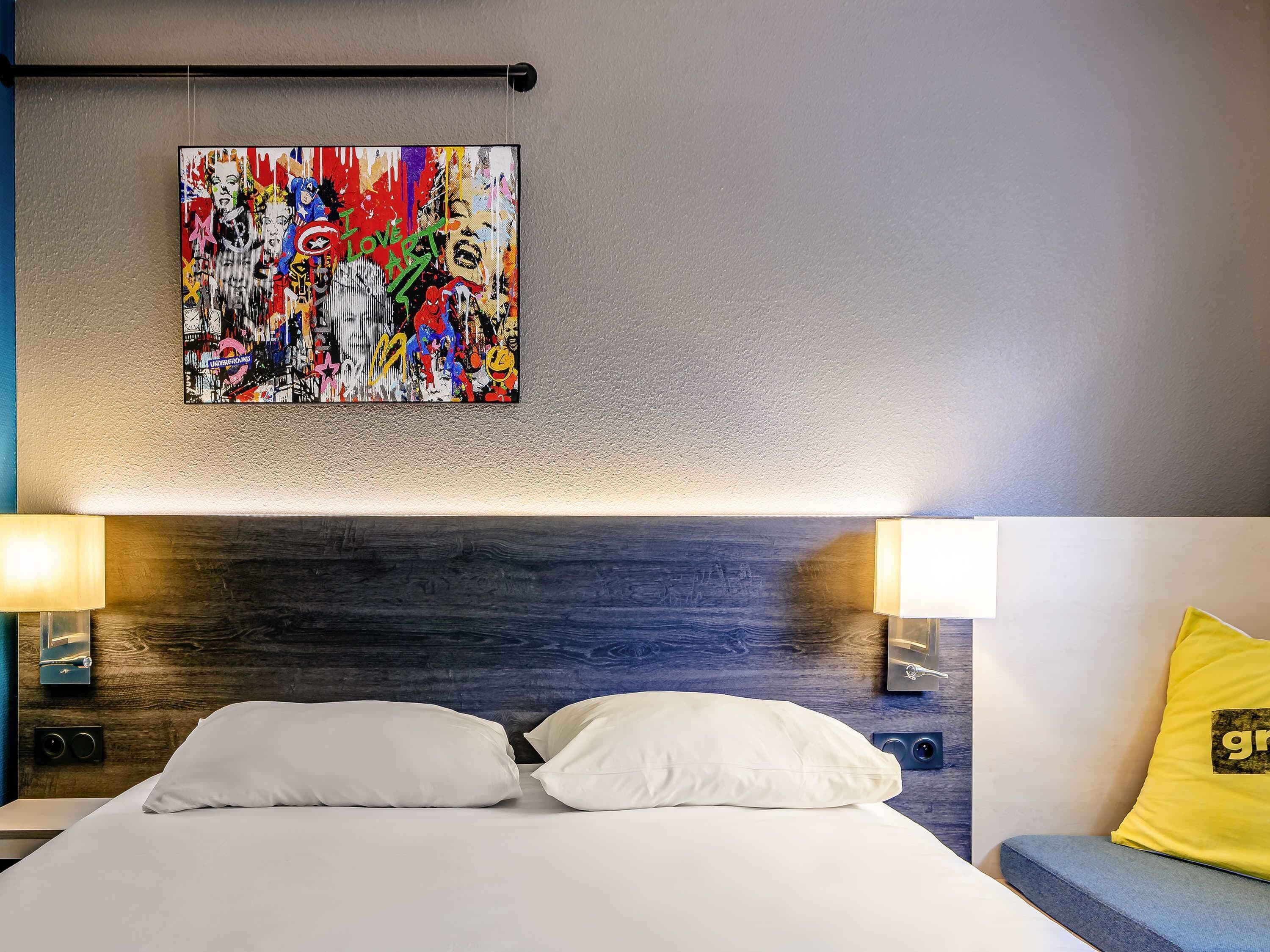 Ein Hotelzimmer mit einem Bett, weißen Kissen, einem farbenfrohen abstrakten Gemälde an der Wand und einem gelben Kissen auf der Couch.