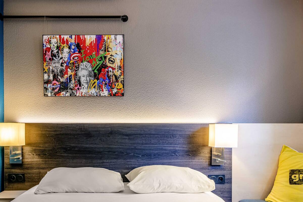Une chambre d’hôtel avec un lit, des oreillers blancs, un tableau abstrait aux couleurs vives accroché au mur et un coussin jaune sur le canapé.