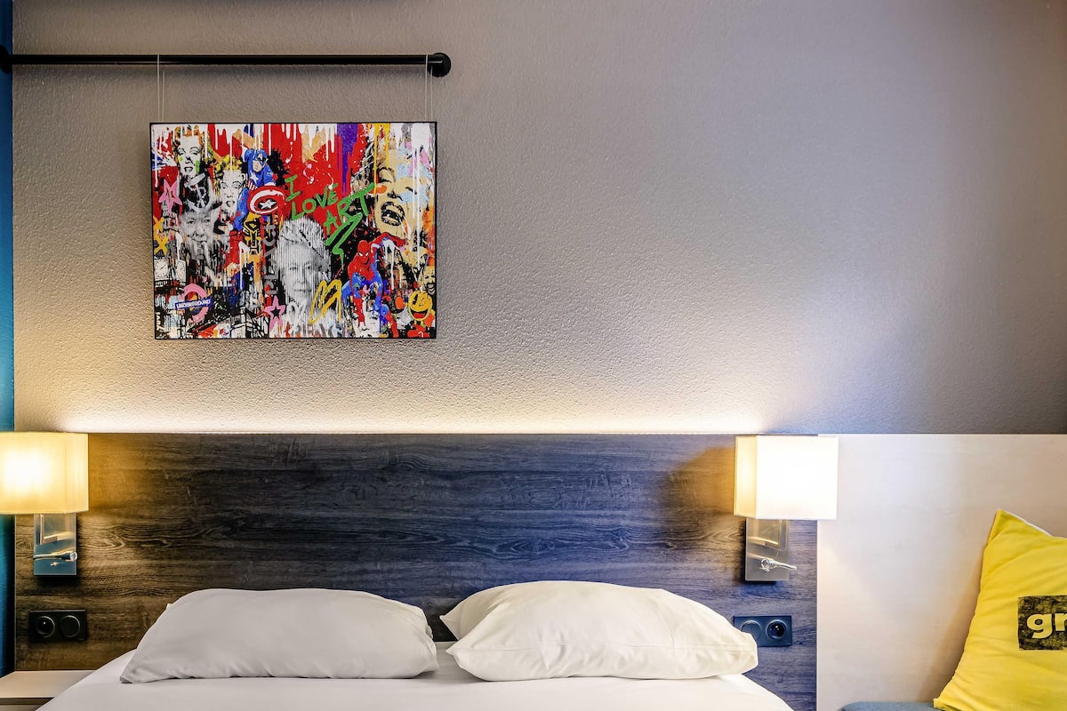 Ein Hotelzimmer mit einem Bett, weiĂen Kissen, einem farbenfrohen abstrakten GemĂ€lde an der Wand und einem gelben Kissen auf der Couch.
