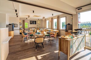 Een ruime eethoek met meerdere tafels en stoelen, een houten toonbank met een tafelkleed met patroon, en grote ramen die natuurlijk licht binnenlaten.