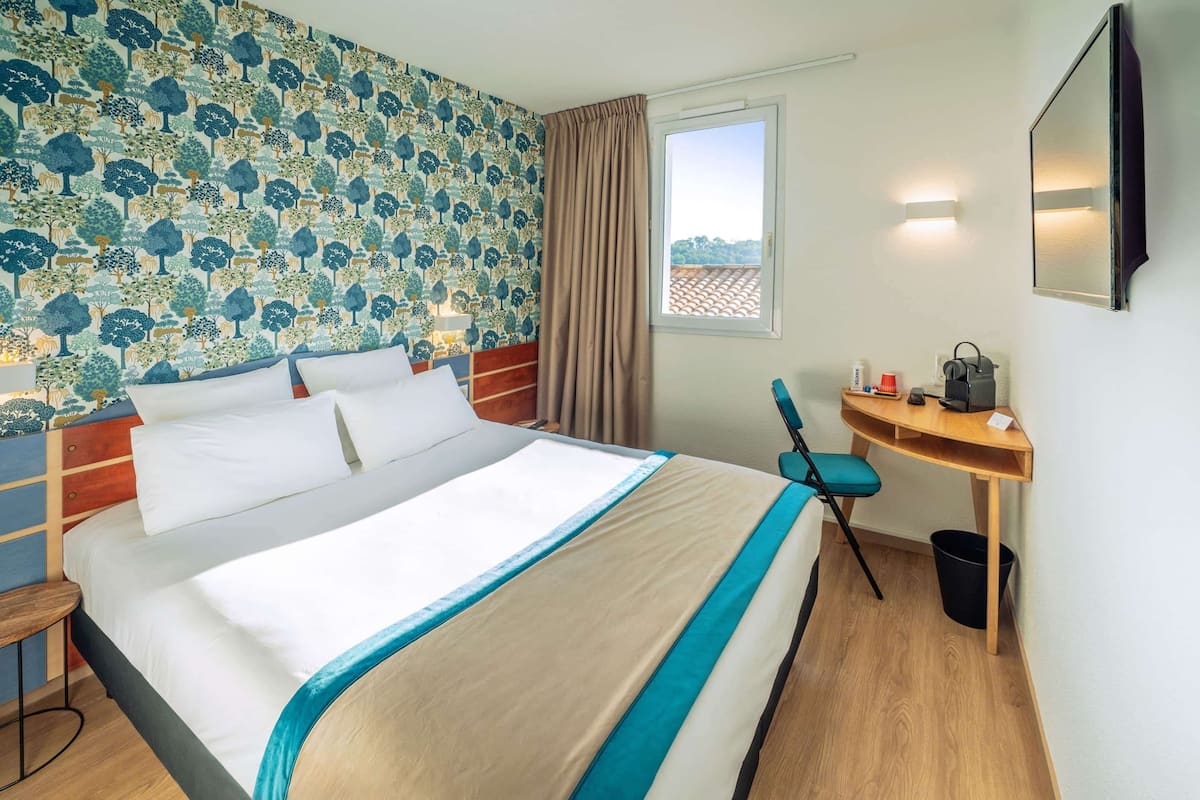 A hotel room with a large bed, a desk with a chair, a window with curtains, and a wall with a patterned wallpaper.