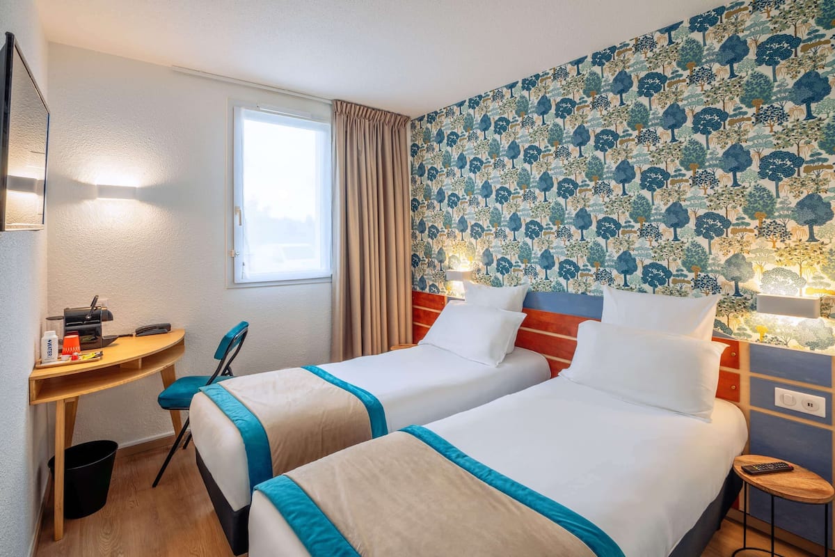 A hotel room with two beds, a desk, a chair, a small table, and a nightstand with a lamp.
