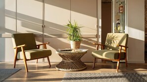 Una habitación con dos sillones verdes, una mesa de centro trenzada con una planta en maceta, y un aparador de vidrio con botellas.