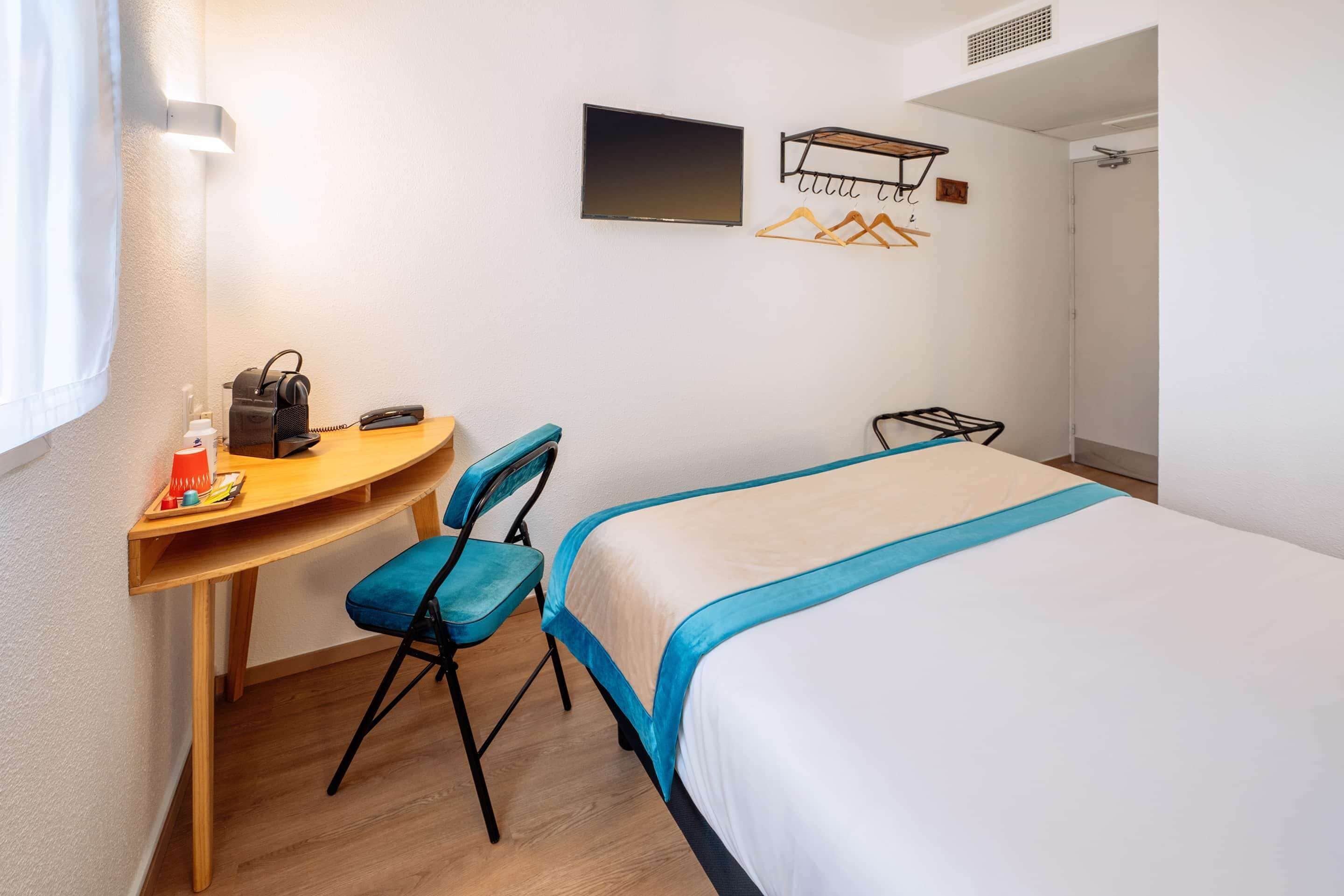 A hotel room with a bed, a desk, a chair, a TV, and a coat rack.