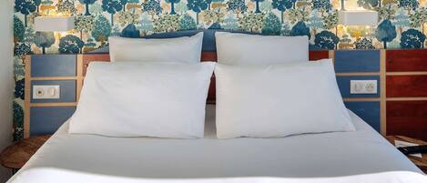 Una cama con ropa de cama blanca y dos almohadas, una mesita de noche con una lámpara, y una pared con un estampado de árbol.