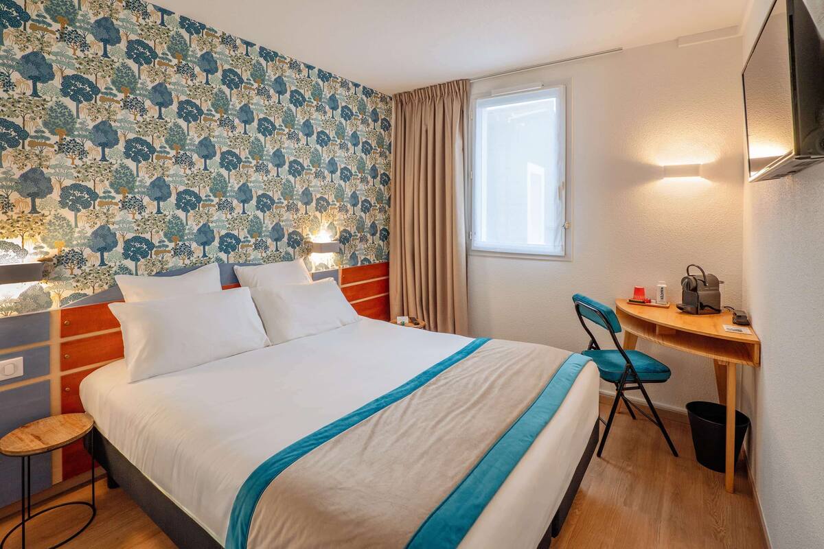 A hotel room with a large bed, a desk with a chair, a small stool, a wall-mounted TV, and a window with curtains.