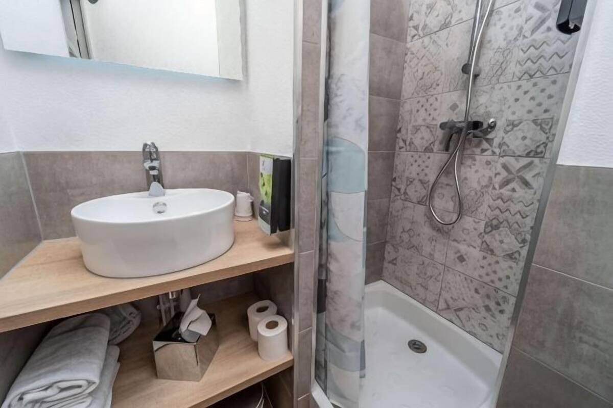 A bathroom with a sink, toilet paper, a shower, and a mirror.
