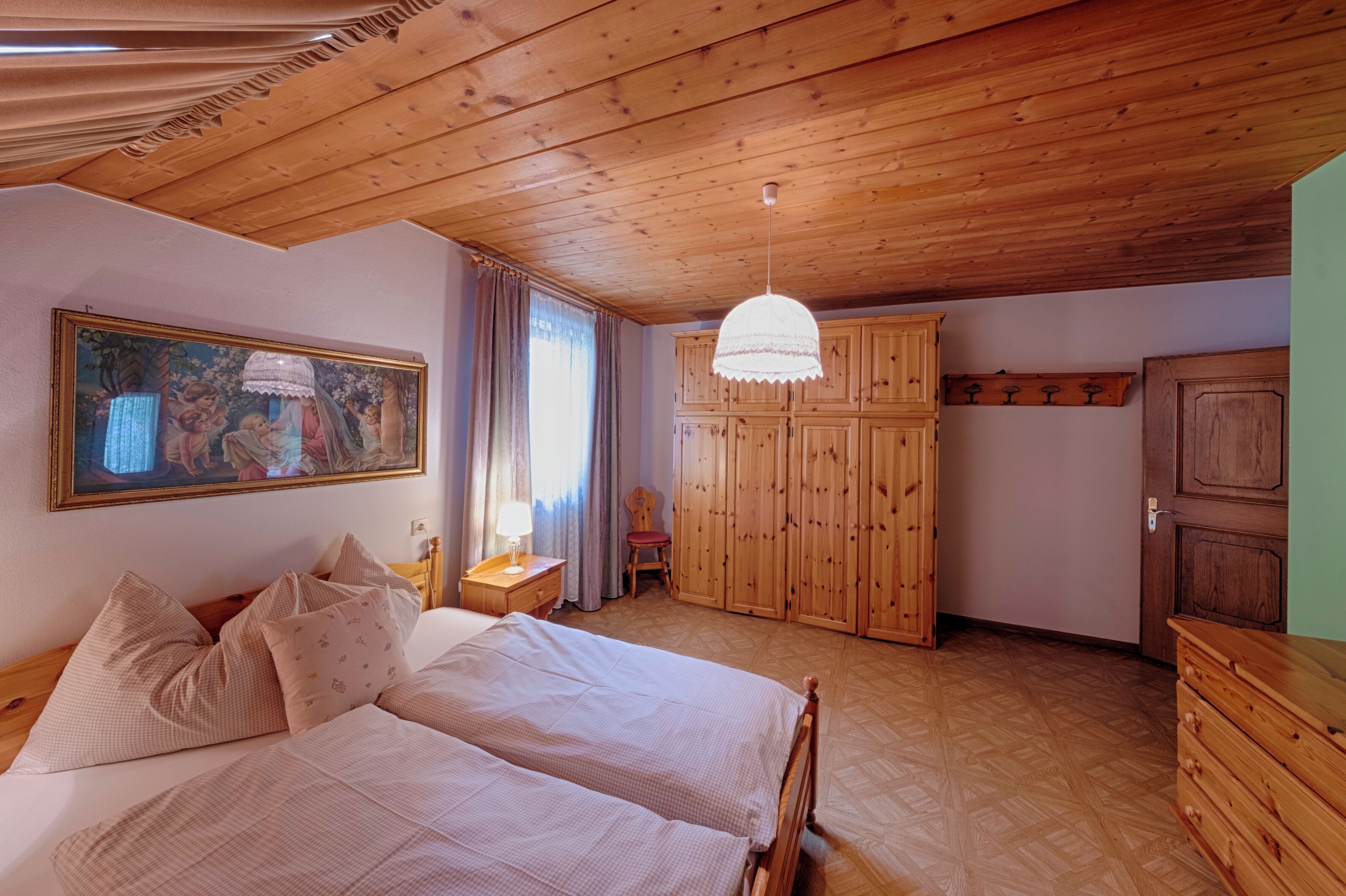 A bedroom with a wooden ceiling, a bed with multiple pillows, a nightstand, a wooden wardrobe, and a painting on the wall.
