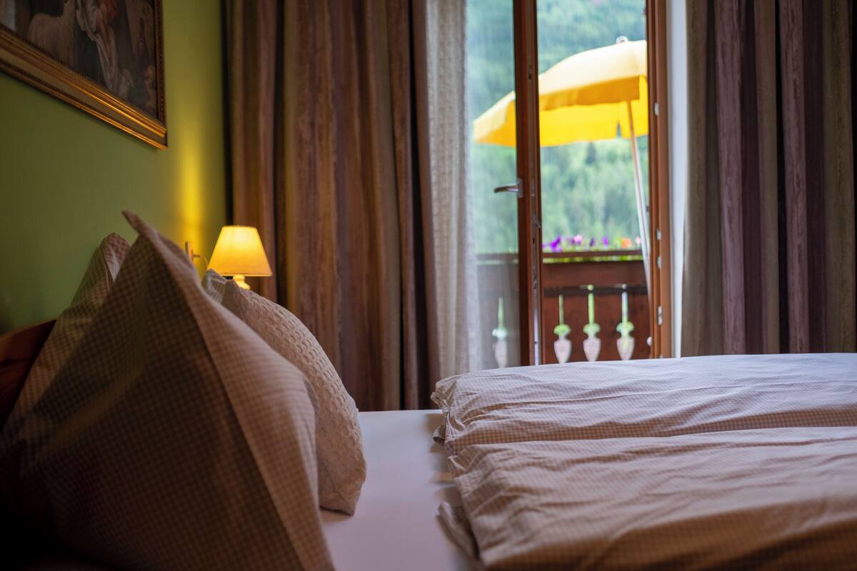 A hotel room with a bed, a lamp, a window with curtains, and a view of greenery outside.