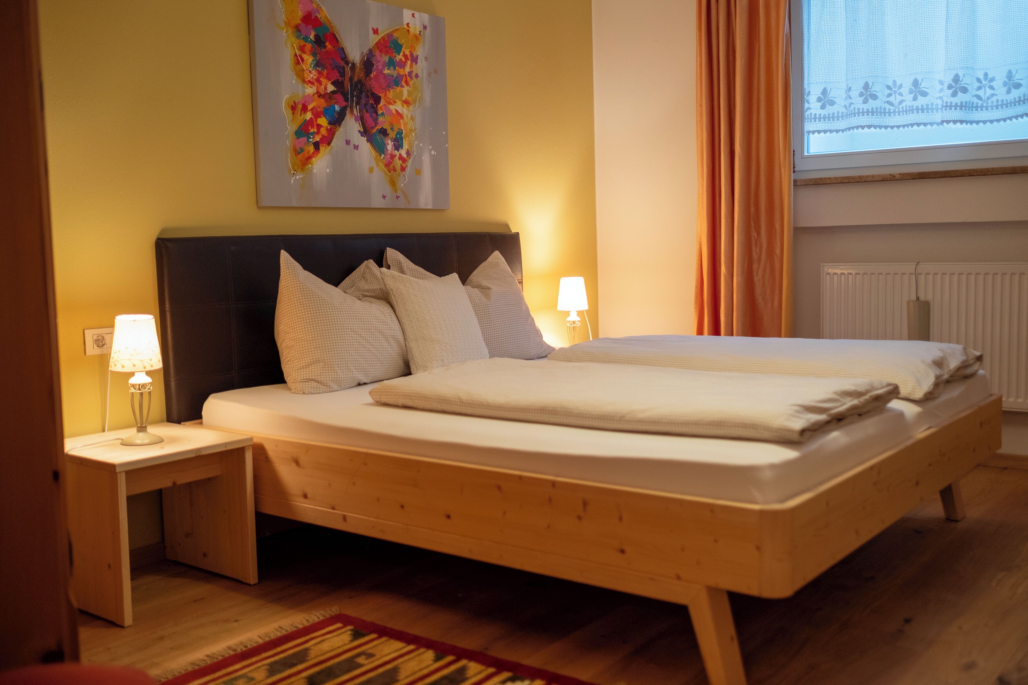 Un dormitorio con una cama de madera, dos mesitas de noche con lámparas, una obra de arte con mariposas coloridas en la pared y una ventana con cortinas.