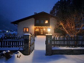 Una casa innevata con un cancello in pietra, una recinzione in legno e un ingresso illuminato.