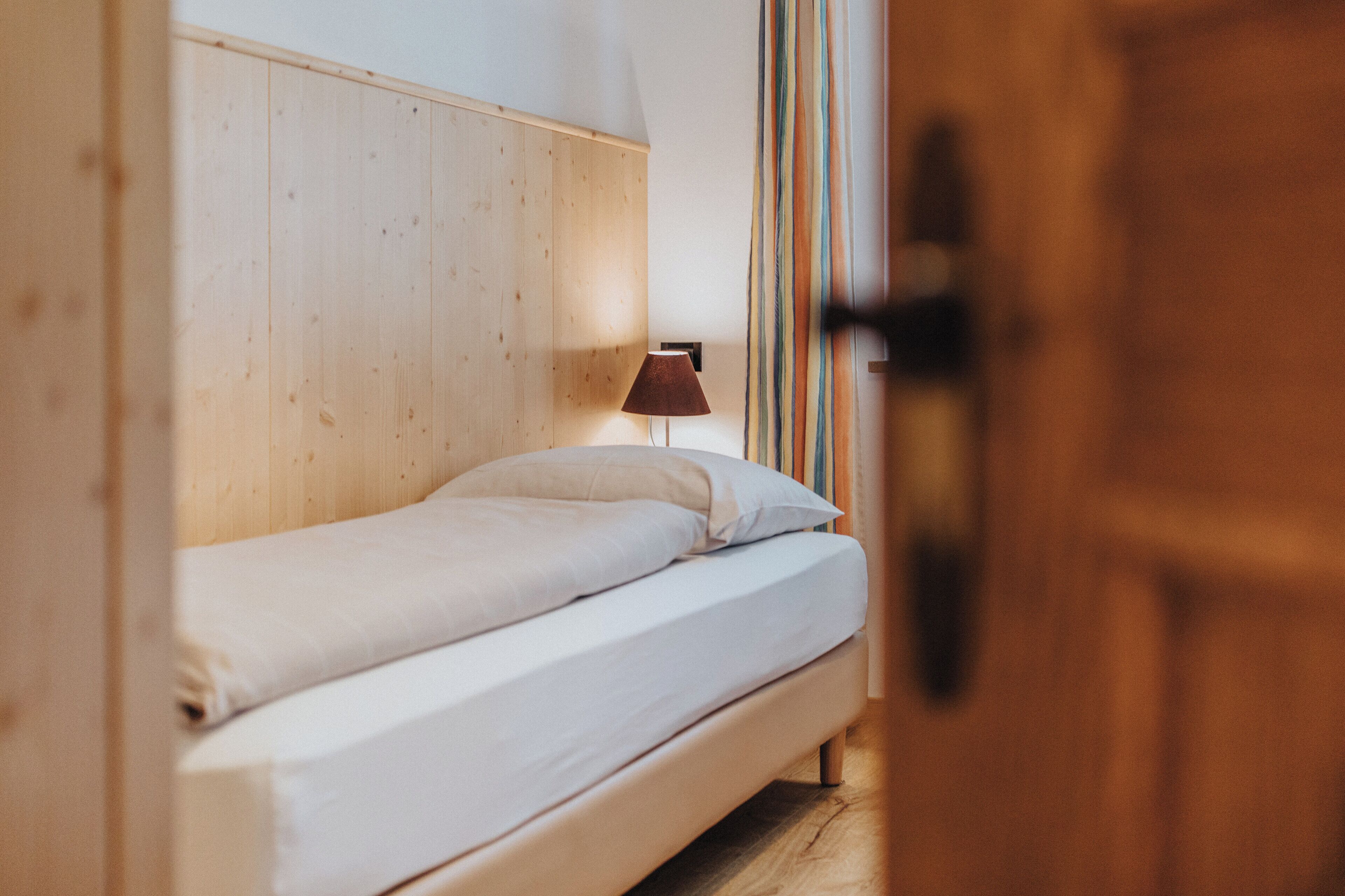 A single bed with a wooden headboard, a bedside lamp, and a striped curtain partially visible.