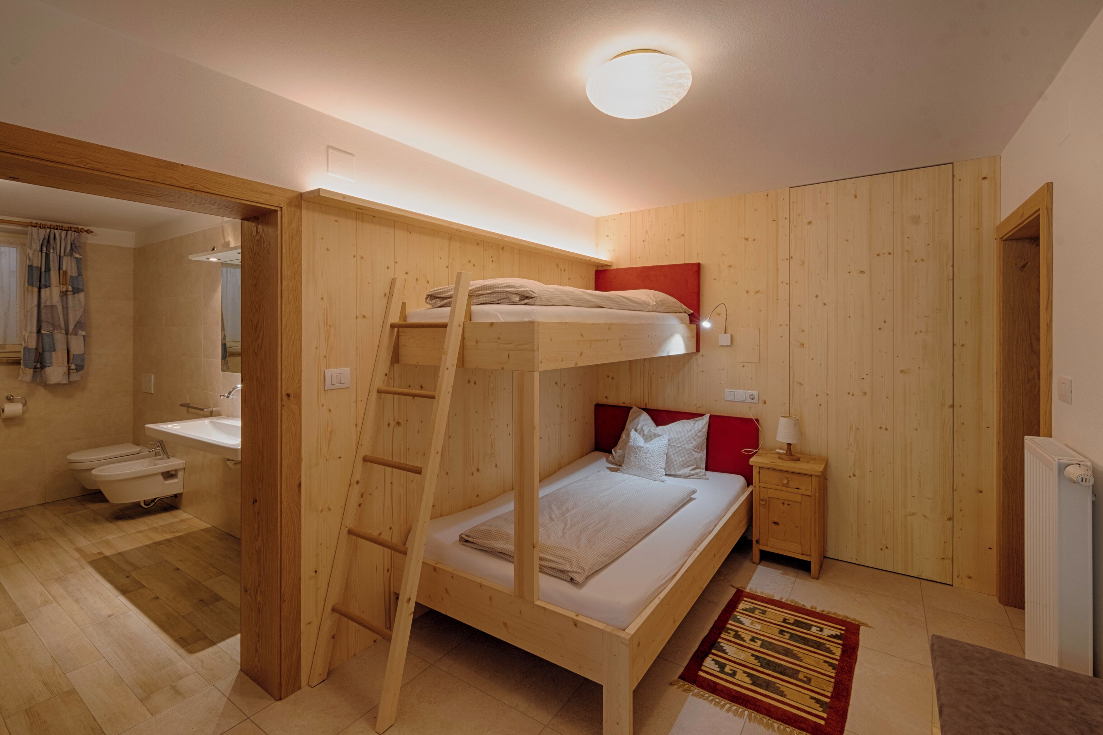 A compact room with a bunk bed, a wooden wardrobe, a small bedside table with a lamp, and a radiator.