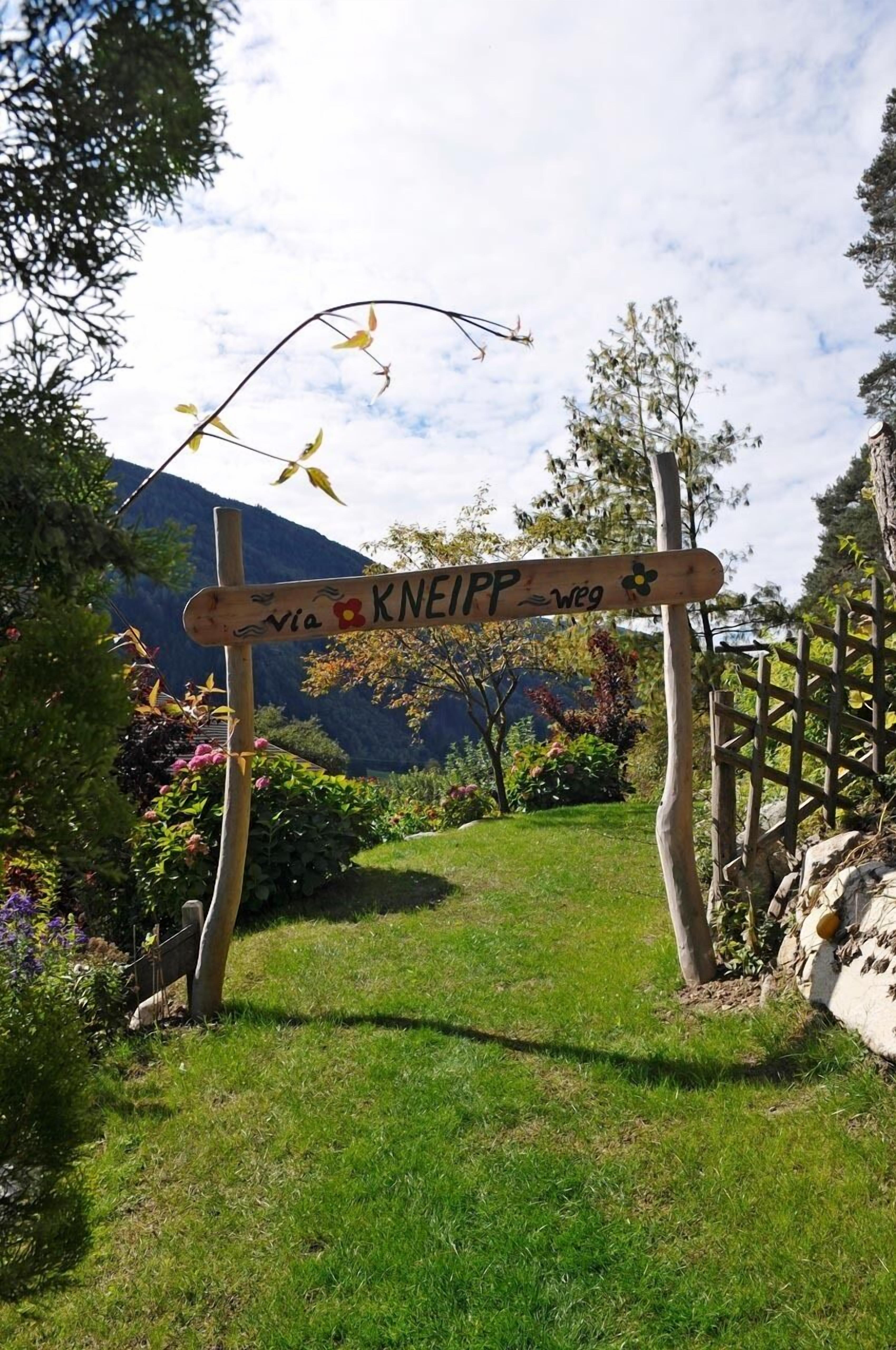 Un cartello in legno con la scritta 'KNEIPP via' e un simbolo di fiore, immerso nel verde con una montagna sullo sfondo.