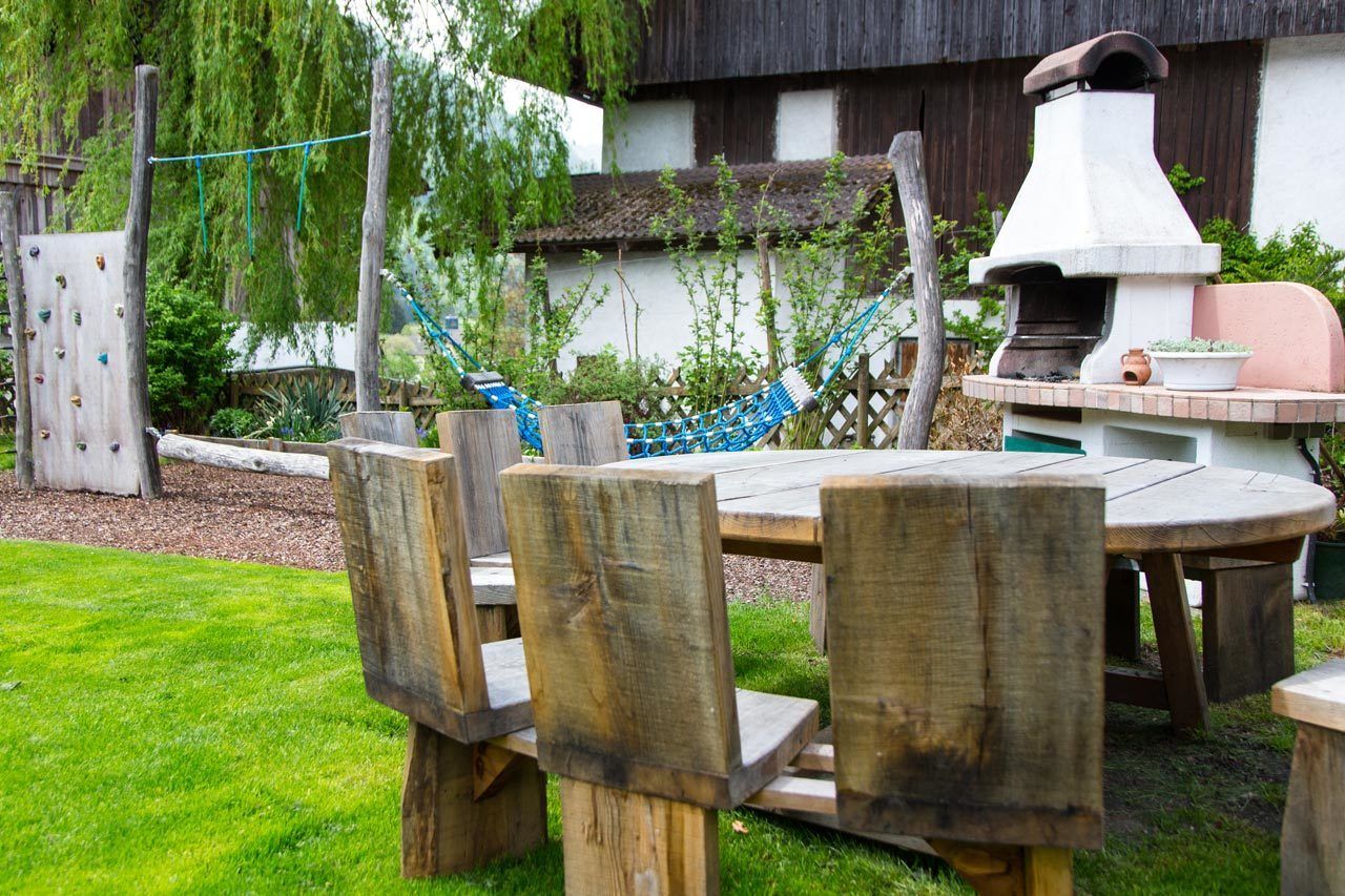 A wooden outdoor dining area with a round table and benches, a hammock, and a stone oven.