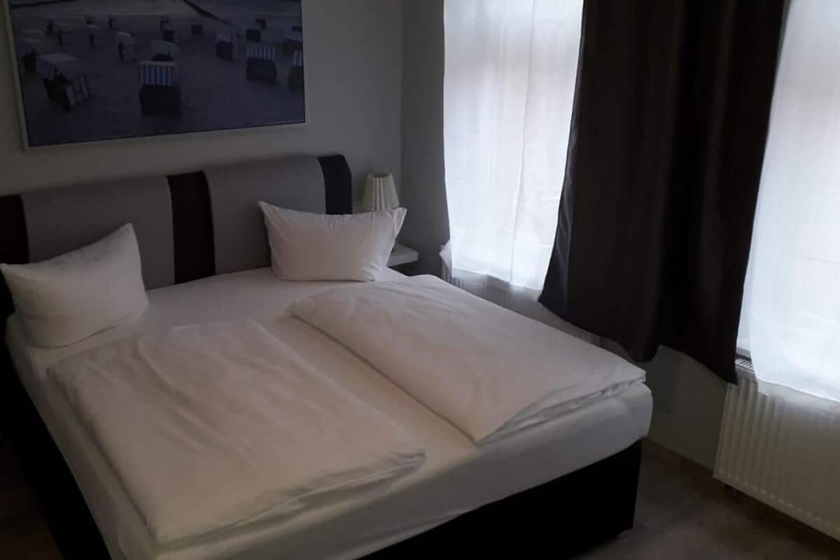 Ein ordentlich bezogenes Bett mit weißen Leinen, ein Kopfteil mit dunklen, horizontalen Streifen und ein Nachttisch mit einer Lampe.