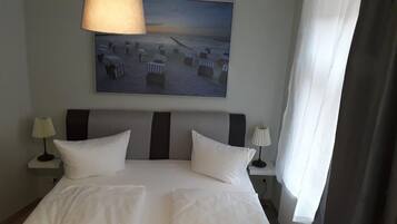 Una camera d'albergo con un letto grande, due lampade ai comodini, un quadro con sedie da spiaggia e una finestra con tende.
