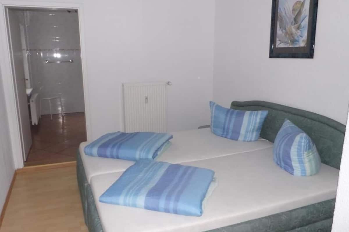 Ein kleines Zimmer mit einem Einzelbett, einem grünen Kopfteil, zwei blau gestreiften Kissen und einem gerahmten Bild an der Wand.